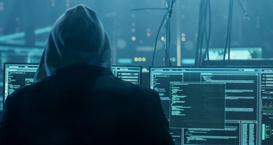 A hacker whose identity is hidden by a hooded top looking at multiple screens